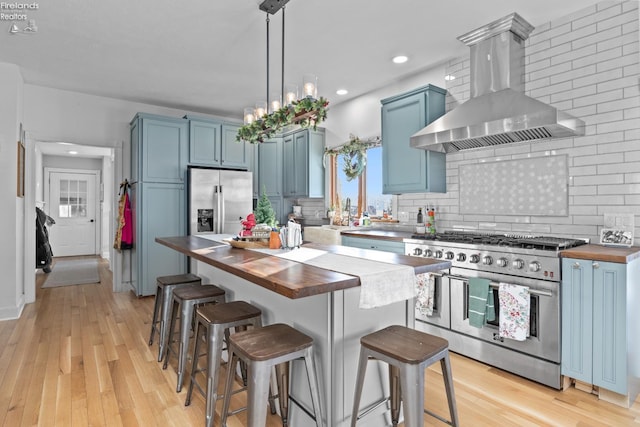 kitchen with butcher block counters, blue cabinets, range hood, and appliances with stainless steel finishes