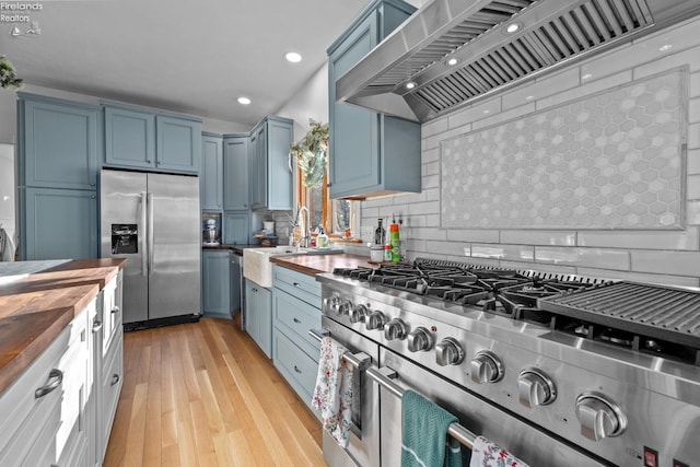 kitchen with butcher block countertops, backsplash, light hardwood / wood-style floors, stainless steel appliances, and wall chimney range hood