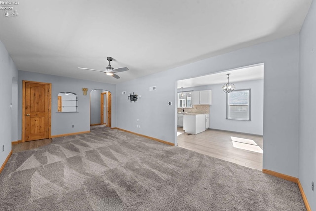 unfurnished living room featuring baseboards, ceiling fan with notable chandelier, arched walkways, and light colored carpet