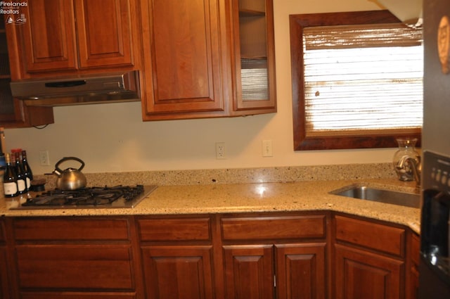 kitchen with glass insert cabinets, light stone countertops, under cabinet range hood, stainless steel gas stovetop, and a sink