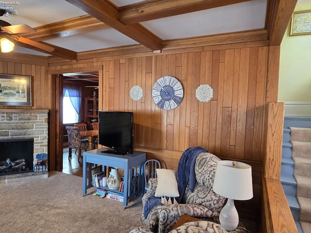 carpeted living room with wooden walls, a ceiling fan, stairway, beamed ceiling, and a stone fireplace