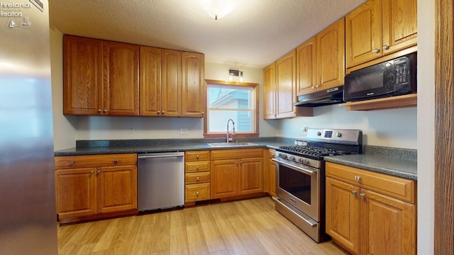 kitchen with dark countertops, stainless steel appliances, light wood-type flooring, under cabinet range hood, and a sink