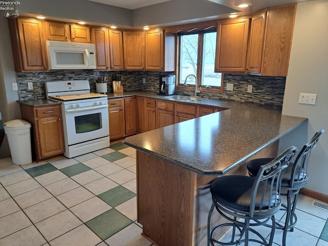 kitchen with a breakfast bar area, dark countertops, a sink, white appliances, and a peninsula