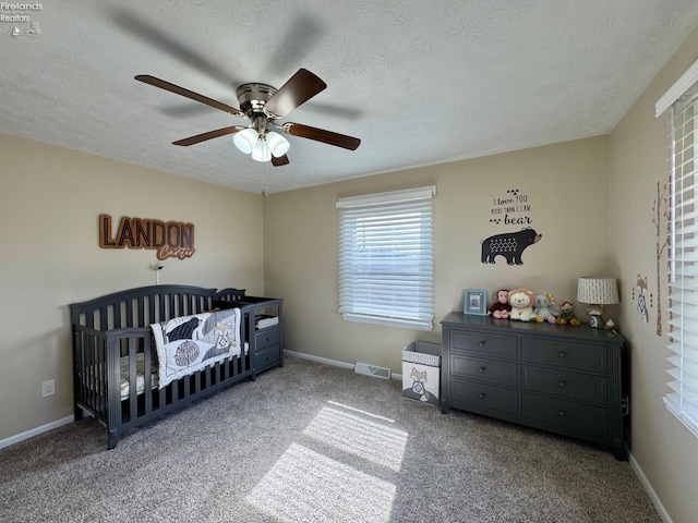 carpeted bedroom featuring a crib, baseboards, visible vents, a ceiling fan, and a textured ceiling