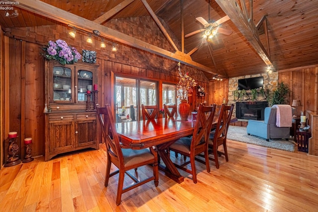 dining room with light wood-style floors, wood ceiling, wood walls, a stone fireplace, and beamed ceiling