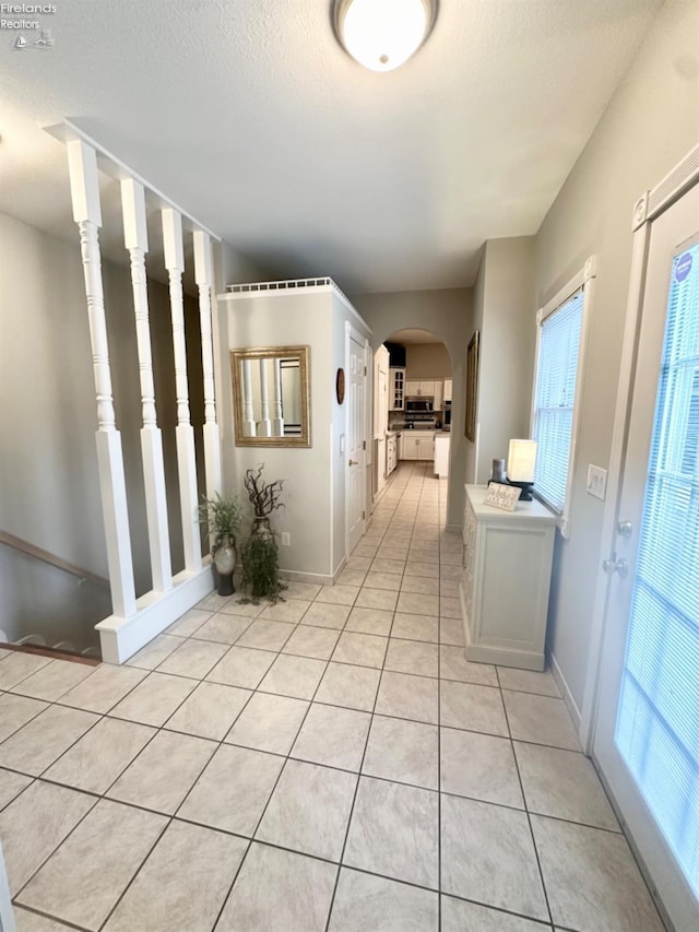 hall with arched walkways, a textured ceiling, baseboards, and light tile patterned floors