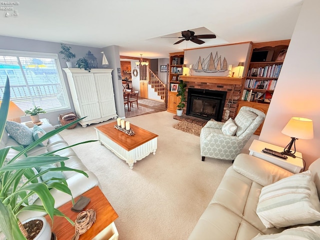 living area featuring stairway, a fireplace, carpet flooring, and ceiling fan with notable chandelier