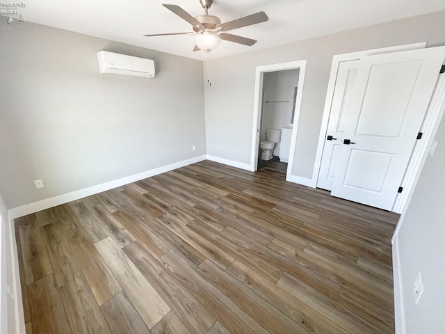 unfurnished bedroom with an AC wall unit, a ceiling fan, ensuite bathroom, wood finished floors, and baseboards