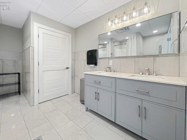 bathroom with a sink, a drop ceiling, tile walls, double vanity, and walk in shower