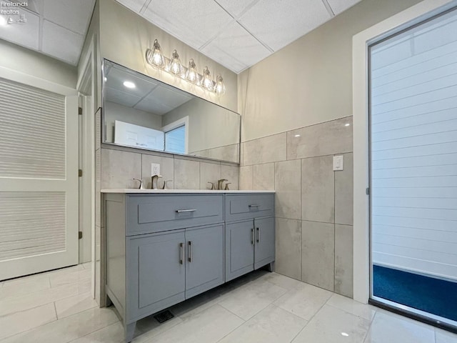 full bath with double vanity, a sink, a paneled ceiling, tile walls, and tile patterned floors