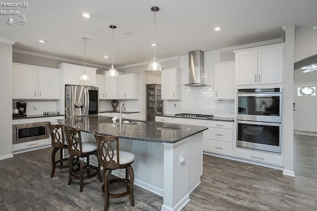 kitchen with dark wood-style floors, a kitchen island with sink, a sink, appliances with stainless steel finishes, and wall chimney range hood