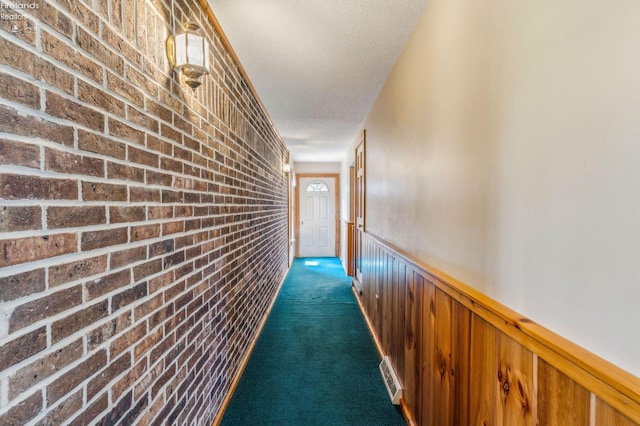 hall with a wainscoted wall, a textured ceiling, visible vents, brick wall, and dark carpet
