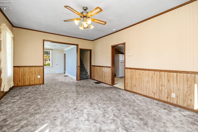 carpeted empty room featuring visible vents, ornamental molding, stairs, a wainscoted wall, and a ceiling fan