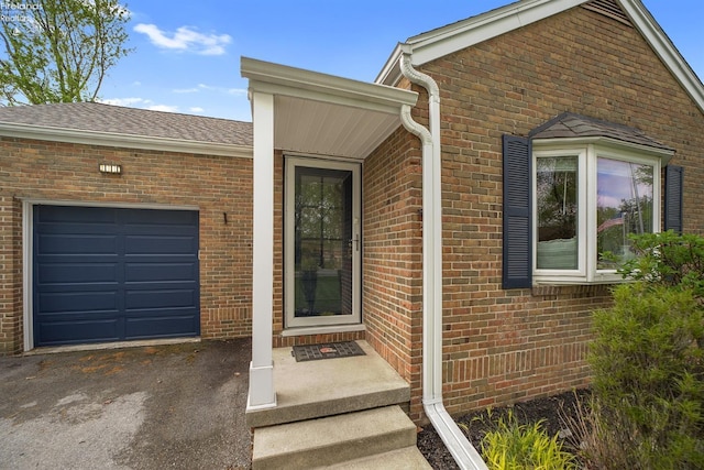 doorway to property with an attached garage, a shingled roof, driveway, and brick siding
