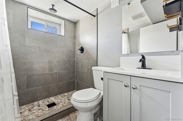 bathroom with a textured ceiling, toilet, vanity, visible vents, and a tile shower