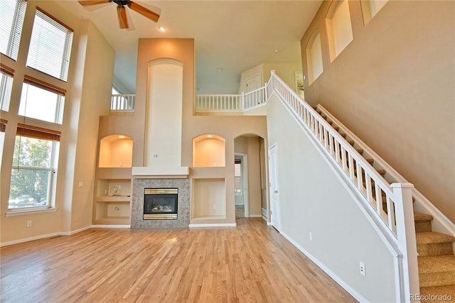 unfurnished living room with light hardwood / wood-style flooring, ceiling fan, built in features, a towering ceiling, and a tiled fireplace