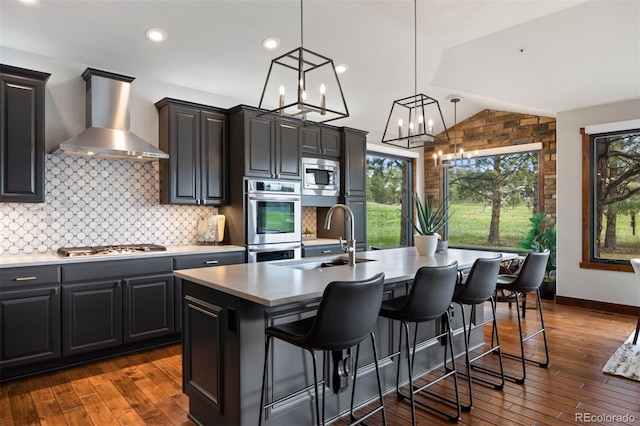 kitchen featuring wall chimney range hood, a sink, stainless steel appliances, a kitchen island with sink, and tasteful backsplash