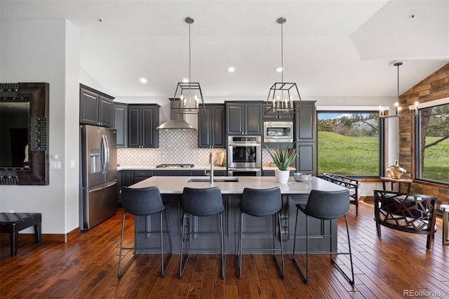 kitchen with appliances with stainless steel finishes, vaulted ceiling, light countertops, wall chimney range hood, and a center island with sink