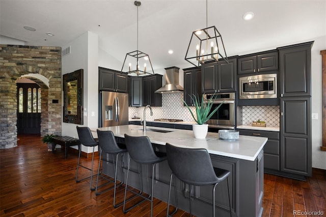 kitchen featuring stainless steel appliances, wall chimney exhaust hood, light countertops, dark wood-style flooring, and a center island with sink