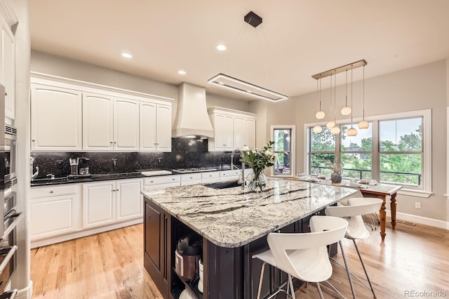 714 Skywalker Pt, Lafayette, CO 80026 - kitchen featuring custom range hood, white cabinetry, light wood-type flooring, backsplash, and light stone counters
