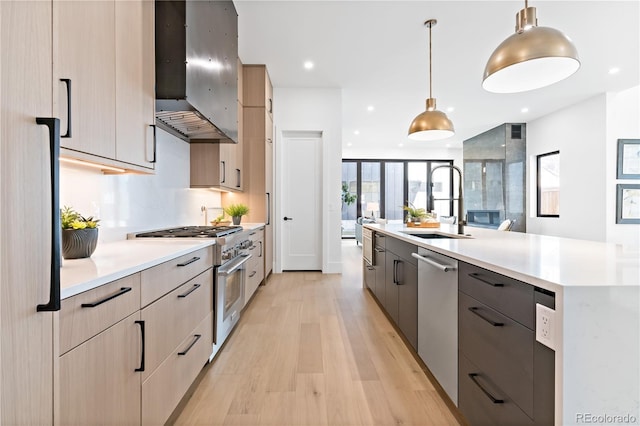 kitchen featuring appliances with stainless steel finishes, light hardwood / wood-style floors, wall chimney exhaust hood, light brown cabinets, and decorative light fixtures