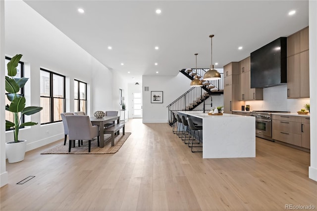 kitchen featuring high end stove, pendant lighting, light hardwood / wood-style flooring, a center island with sink, and wall chimney range hood