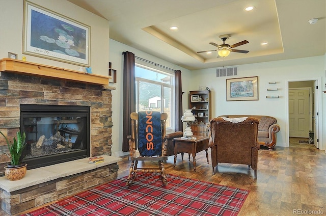 sitting room featuring ceiling fan, a stone fireplace, visible vents, wood finished floors, and a raised ceiling