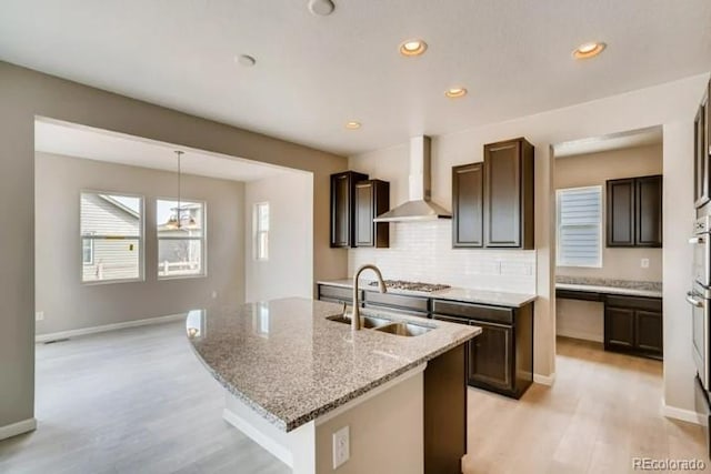 kitchen featuring backsplash, a sink, wall chimney range hood, dark brown cabinets, and light stone countertops