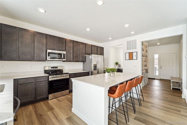 kitchen with appliances with stainless steel finishes, dark brown cabinets, a kitchen island, a kitchen bar, and light wood-type flooring