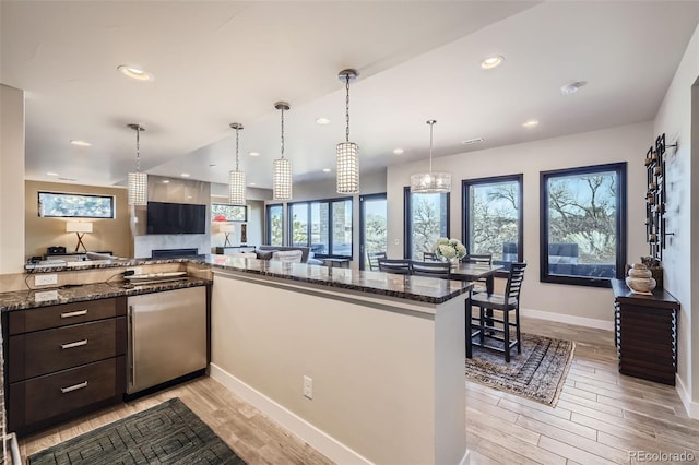 kitchen featuring kitchen peninsula, dark stone counters, dark brown cabinetry, fridge, and decorative light fixtures