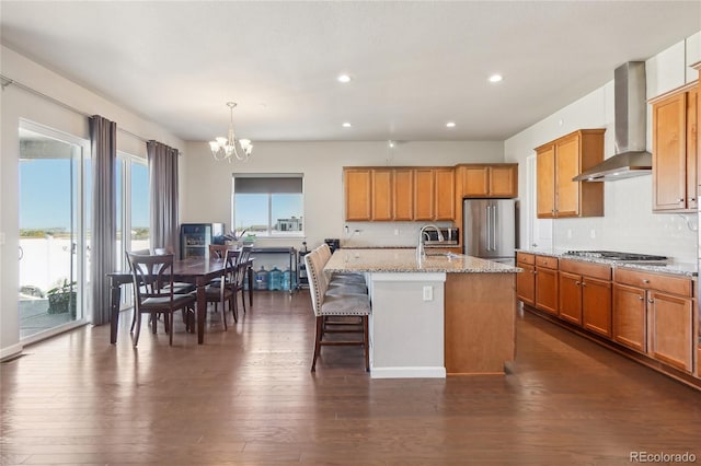 kitchen with pendant lighting, a center island with sink, wall chimney range hood, stainless steel appliances, and dark hardwood / wood-style flooring