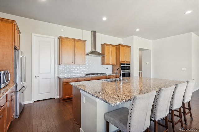 kitchen with light stone counters, a kitchen island with sink, dark wood-type flooring, wall chimney range hood, and stainless steel appliances