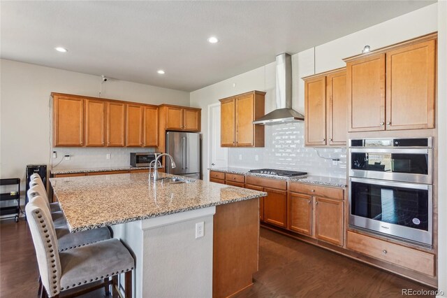 kitchen featuring appliances with stainless steel finishes, wall chimney exhaust hood, dark hardwood / wood-style flooring, and an island with sink