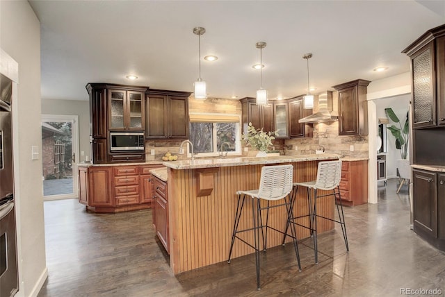 kitchen with wall chimney exhaust hood, appliances with stainless steel finishes, glass insert cabinets, a center island, and pendant lighting