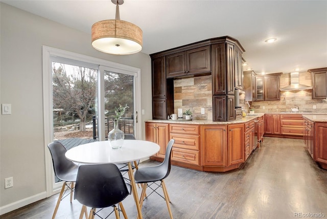 kitchen with brown cabinets, hanging light fixtures, decorative backsplash, wood finished floors, and wall chimney exhaust hood