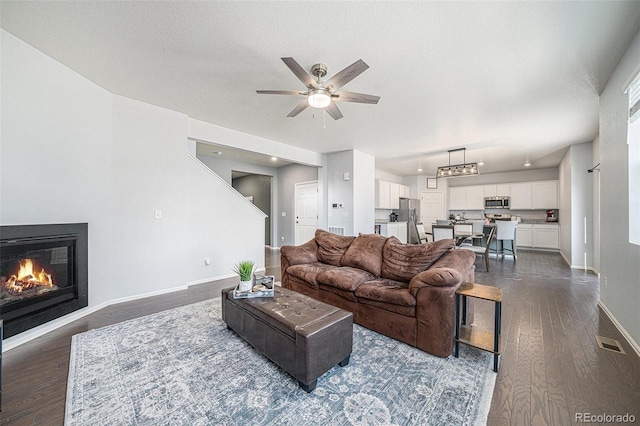 living area featuring dark wood-style flooring, a ceiling fan, a glass covered fireplace, a textured ceiling, and baseboards