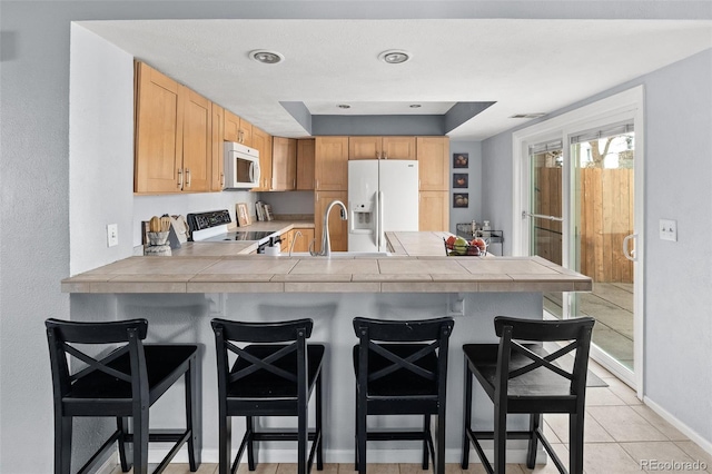 kitchen featuring light tile patterned floors, a peninsula, white appliances, a kitchen breakfast bar, and tile counters