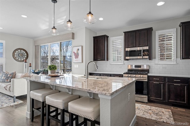 kitchen featuring a kitchen island with sink, sink, stainless steel appliances, and hanging light fixtures