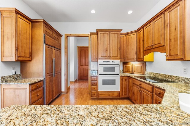 kitchen with black electric cooktop, light wood-type flooring, light stone counters, white double oven, and paneled refrigerator