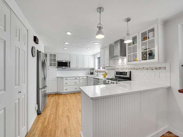 kitchen featuring appliances with stainless steel finishes, a peninsula, wall chimney range hood, white cabinetry, and a sink