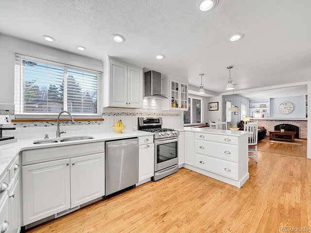 kitchen featuring light wood finished floors, a peninsula, stainless steel appliances, wall chimney range hood, and a sink