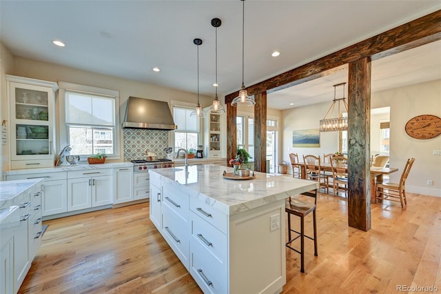 kitchen with light stone countertops, wall chimney exhaust hood, pendant lighting, white cabinets, and an island with sink