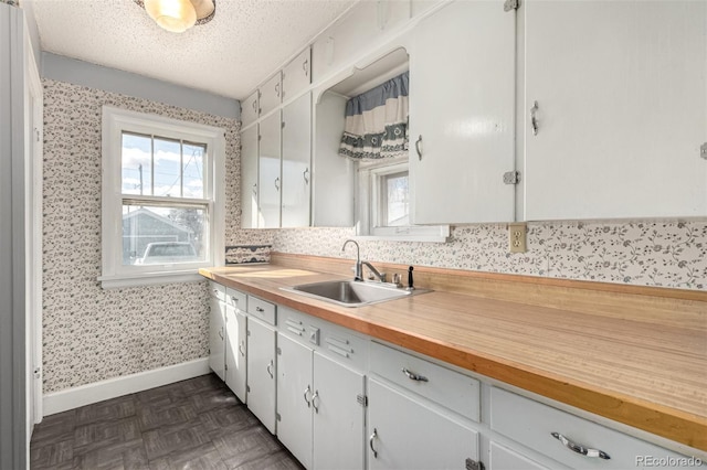 kitchen featuring a sink, a textured ceiling, wood counters, baseboards, and wallpapered walls