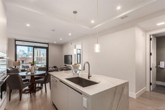 kitchen featuring a center island with sink, sink, white cabinetry, hanging light fixtures, and light wood-type flooring