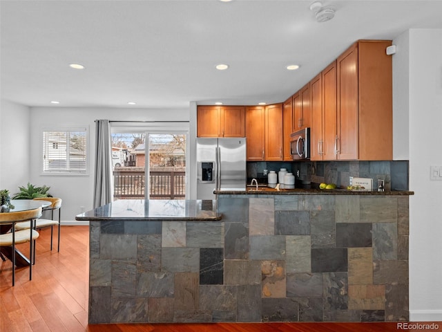 kitchen with stainless steel appliances, light wood finished floors, brown cabinetry, dark stone countertops, and tasteful backsplash