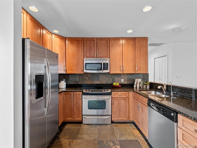 kitchen with backsplash, appliances with stainless steel finishes, stone finish flooring, a sink, and dark stone counters