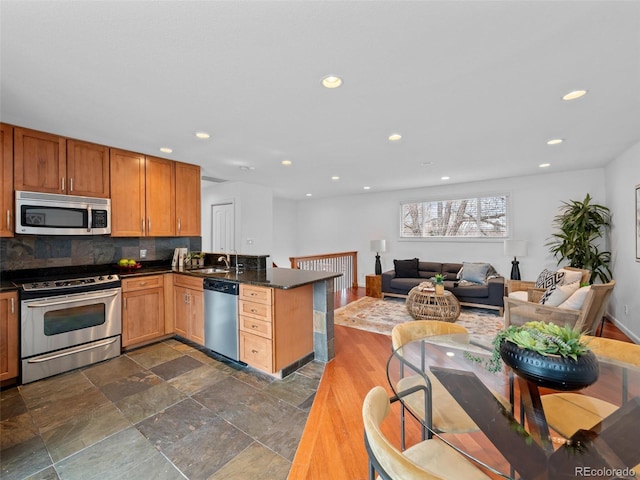 kitchen featuring stainless steel appliances, a peninsula, a sink, backsplash, and dark countertops