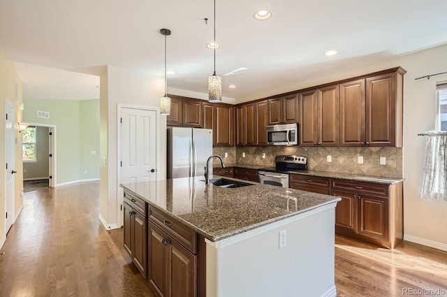 kitchen featuring a kitchen island with sink, appliances with stainless steel finishes, sink, dark stone countertops, and light hardwood / wood-style flooring