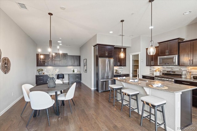 kitchen featuring pendant lighting, dark wood-type flooring, stainless steel appliances, light stone countertops, and an island with sink