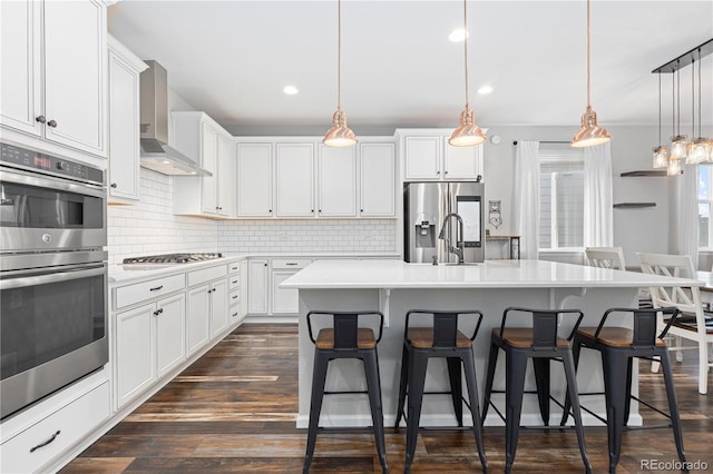 kitchen with wall chimney range hood, appliances with stainless steel finishes, white cabinetry, hanging light fixtures, and an island with sink